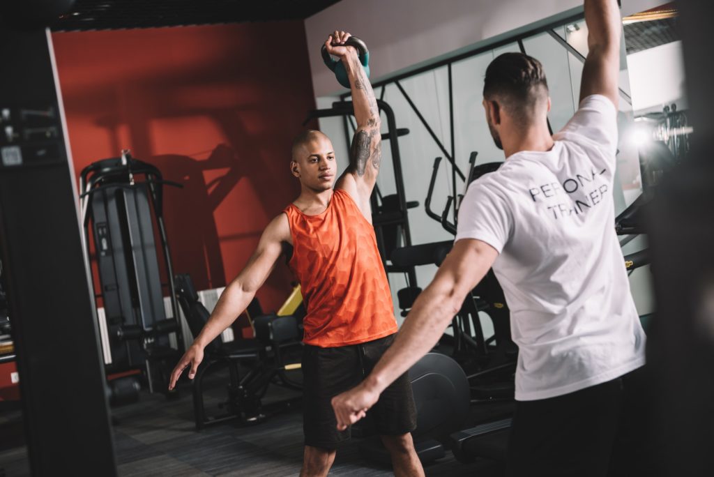 back view of personal trainer lifting weight together with young african american athlele
